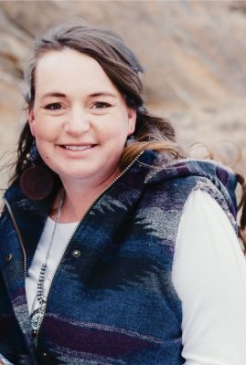 A woman with shoulder-length wavy brown hair wearing a dark blue vest over a white shirt.