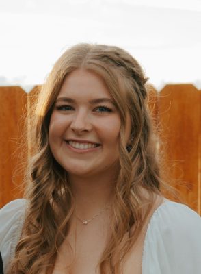 A young woman with long wavy blonde hair wearing an off-white shirt and a necklace.