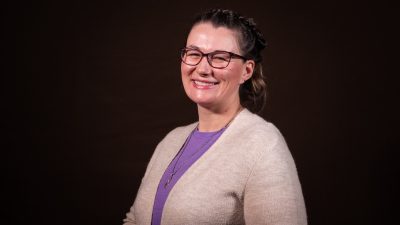 A woman with bobbed brown hair wearing glasses, a purple shirt, and tan cardigan.