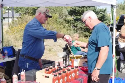 farmers' market vendor squeezes sauce onto a spoon for a customer to try. Both men are standing under a white vendor tent and there is a table with bottles of sauce lined up between them