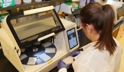 laboratory technician wearing PPE (glasses, coat, gloves) loads samples into a machine to be analyzed