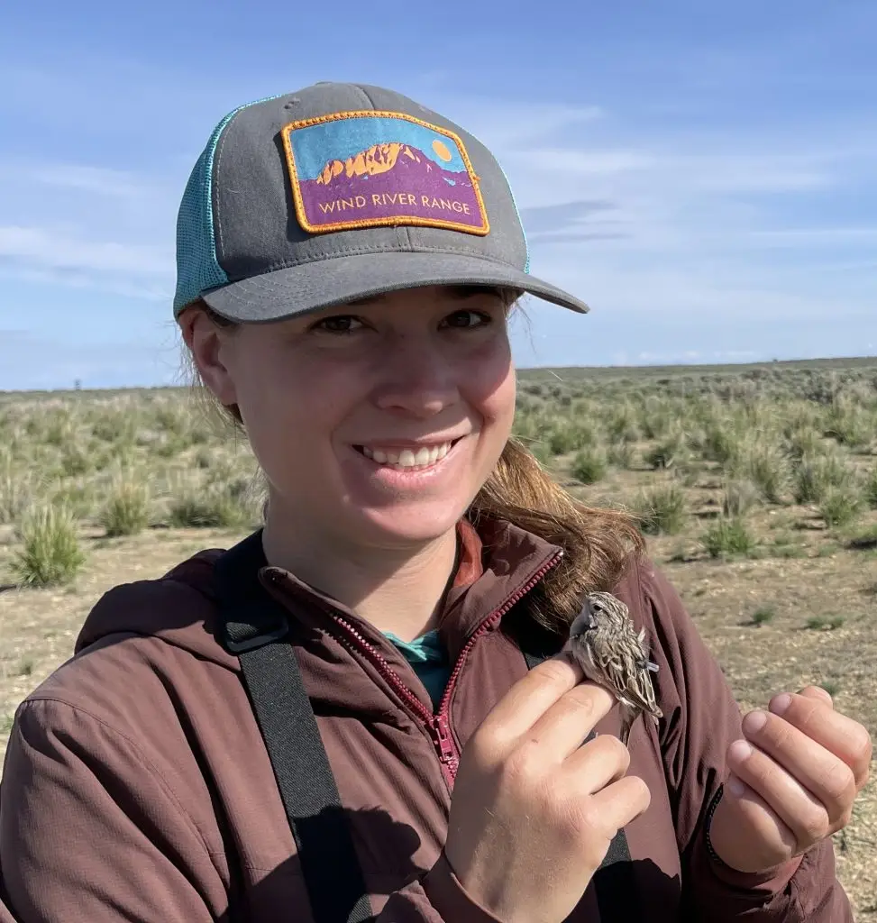 smiling woman wearing a baseball cap holds songbird on two fingers