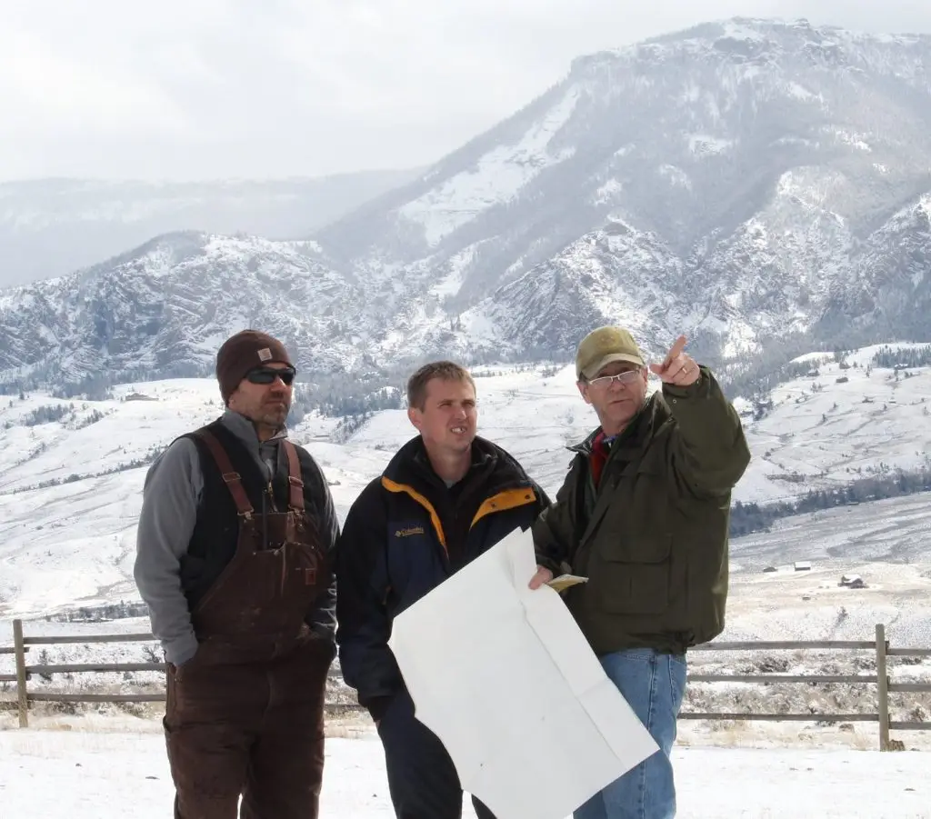 three men, one holding up a pointing finger, look past the camera into the distance with a snowy mountain in the background behind them
