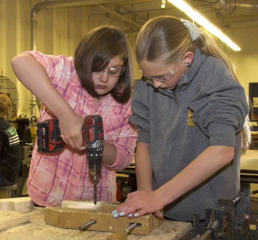 two girls wearing safety glasses work together on a woodworking project, one of them holding a drill and the other stabilizing a piece of wood on the table