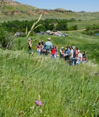 group of people gather around a presenter on a grassy hillside with a cluster of vehicles beside green trees below and a slope with red rock in the background