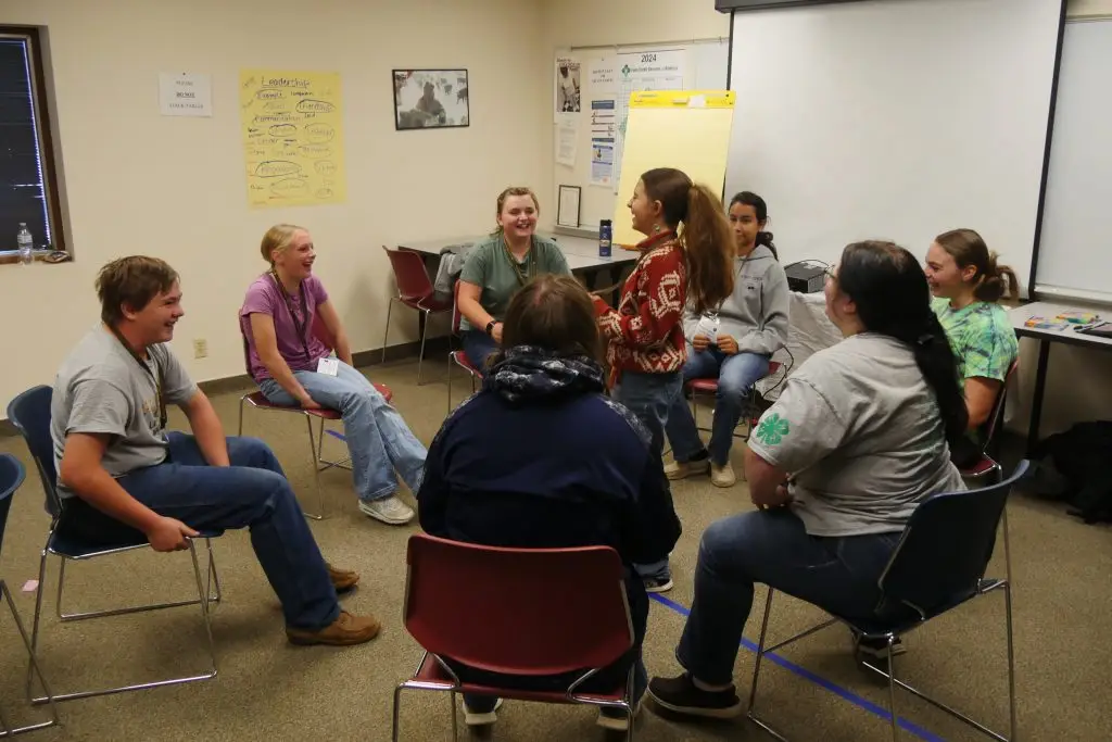 seven young teens sit in chairs around an eighth girl who is standing in the middle of the circle. Everyone is smiling and laughing.
