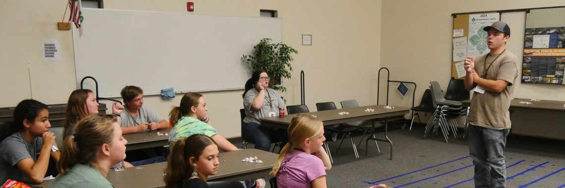 man stands in front of eight 4-H members who are seated at long tables facing toward the presenter