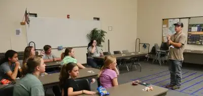 man stands in front of eight 4-H members who are seated at long tables facing toward the presenter