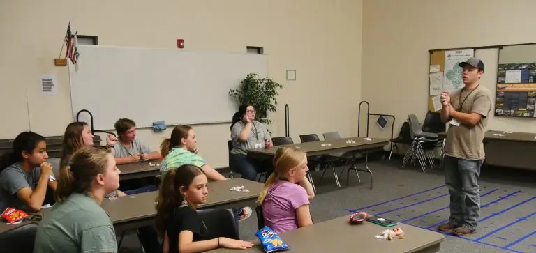 man stands in front of eight 4-H members who are seated at long tables facing toward the presenter
