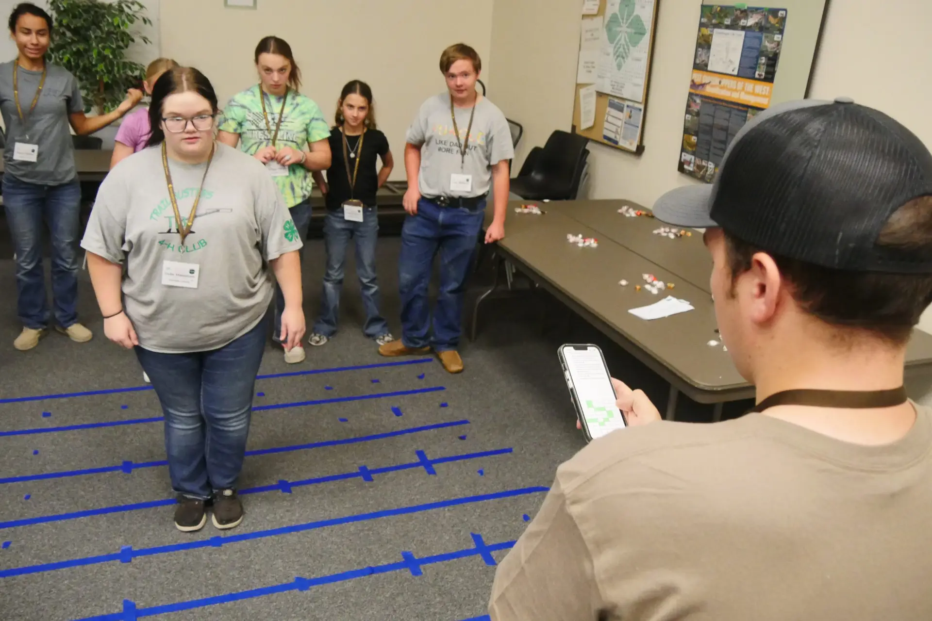 a man in the foreground holding a smart phone faces six 4-H members standing in or behind a series of blue tape strips on the floor