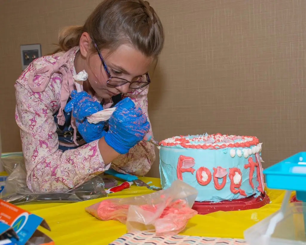 girl wearing frosting-splattered gloves uses a piping bag to frost a cake
