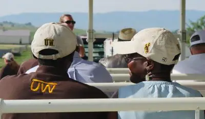 two people wearing University of Wyoming branded caps and clothing face forward, listening to a speaker standing at the front of a metal wagon during a research tour in the fields