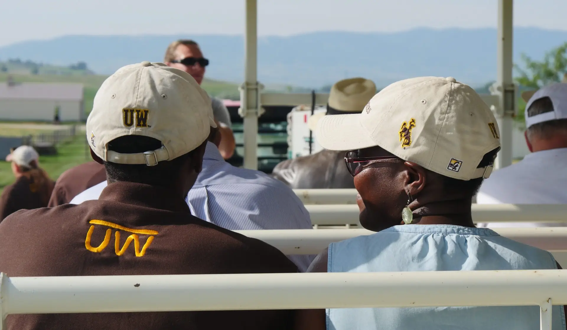 two people wearing University of Wyoming branded caps and clothing face forward, listening to a speaker standing at the front of a metal wagon during a research tour in the fields