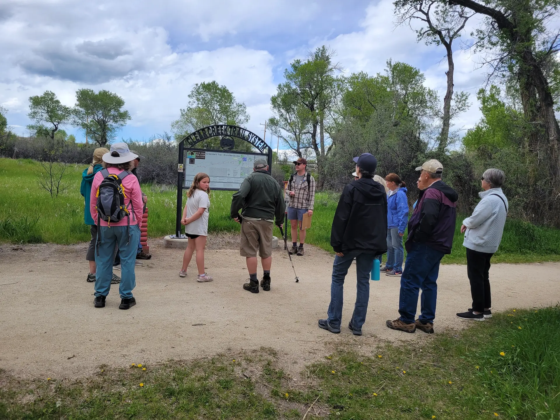 group of people in hiking attire gather around a metal trailhead sign where a man with a clipboard is standing