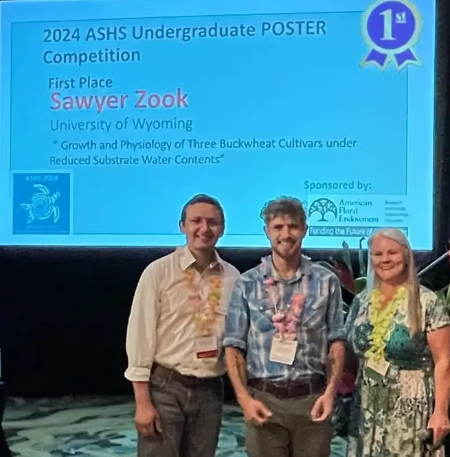 three smiling people stand in front of a screen titled 2024 ASHS Undergraduate Poster Competition with a blue first place ribbon in the top right and Sawyer Zook's name below