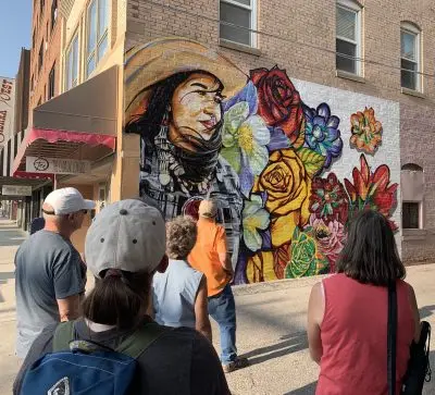 five people facing away from the camera stand on a city street looking up at a mural painted on the side of a brick building depicting a woman in Western clothing beside large flowers