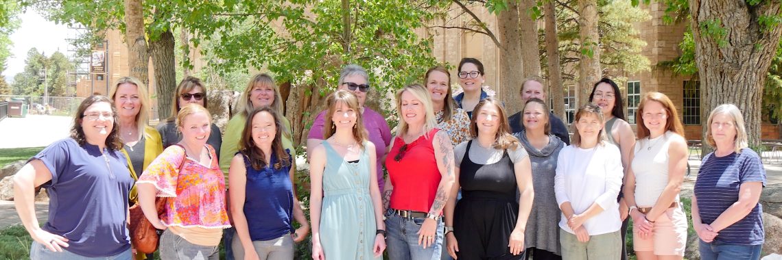 18 smiling women stand together two rows deep under several trees on a college campus