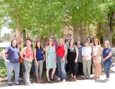18 smiling women stand together two rows deep under several trees on a college campus