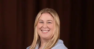 Portrait of a smiling woman with long straight blonde hair, wearing a light blue and white striped top, in front of a dark brown backdrop.