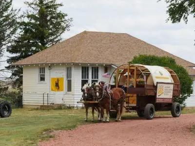 Two horses with a sheep wagon outside a small building.