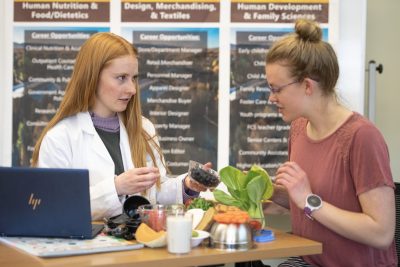 A young woman wearing a labcoat talks with another young woman and shows her a bowl of blueberries. Other healthy foods are spread out between them.