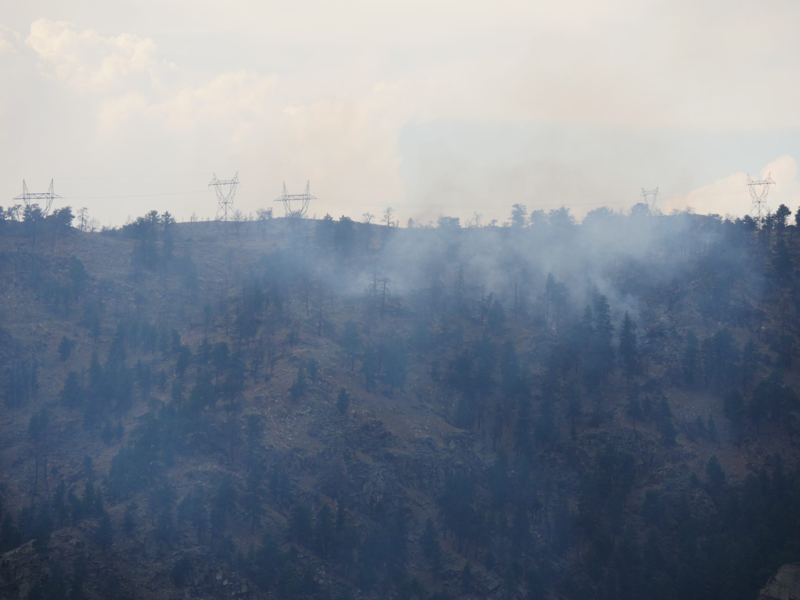 hazy smoke rises from a steep rocky slope scattered with pine trees. At the top of the slope, metal utility infrastructure and lines are visible through the haze.