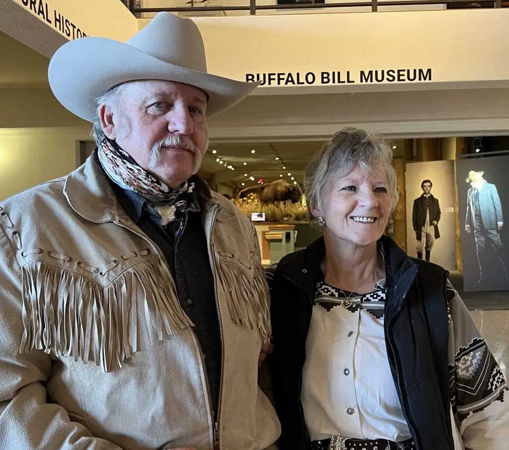 man and woman wearing Western garb stand in front of a museum exhibit with a Buffalo Bill Museum sign on the wall