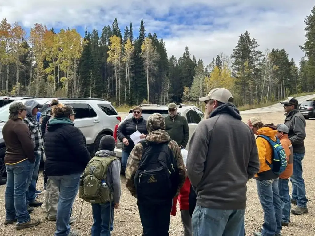 group of people bundled up in outdoor gear stand in a circle around two walk leaders in a parking lot surrounded by trees, including aspens with yellow leaves