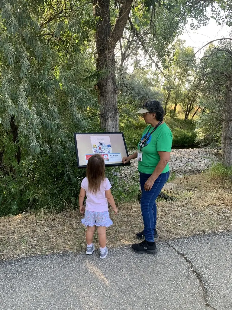 woman and child stand on a paved path in a park and look at a display featuring a picture book