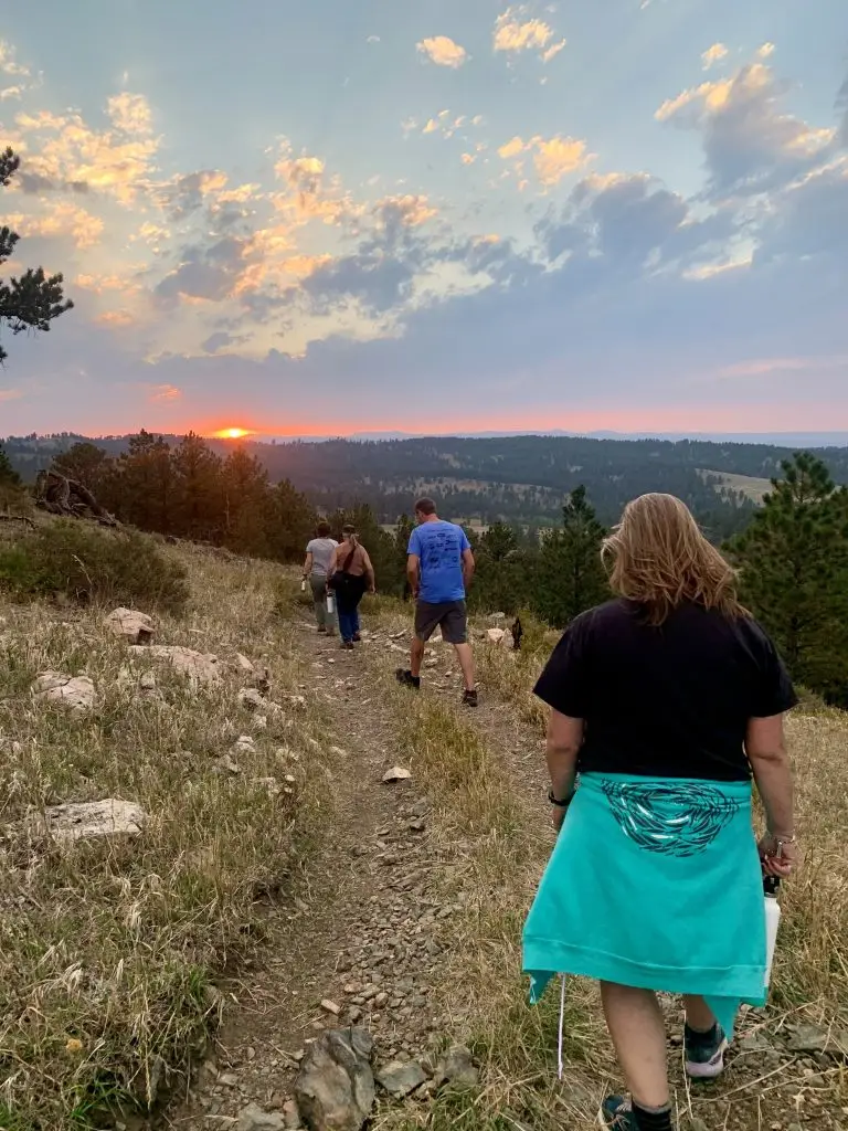 four hikers walk away from the camera down a rocky trail toward the sun setting over the trees