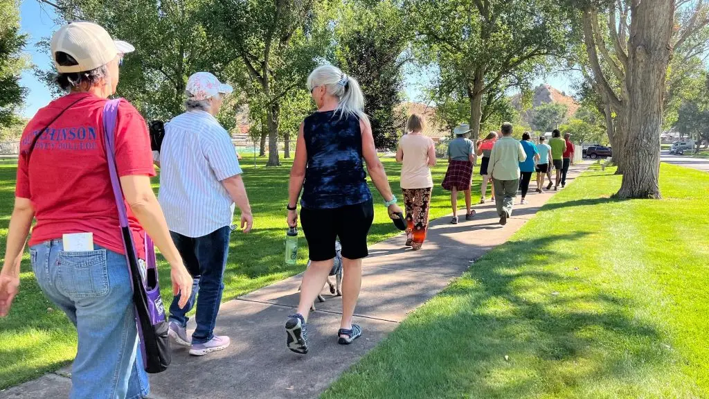 line of people walk down a concrete sidewalk bordered by green grass and trees toward a red rock outcropping
