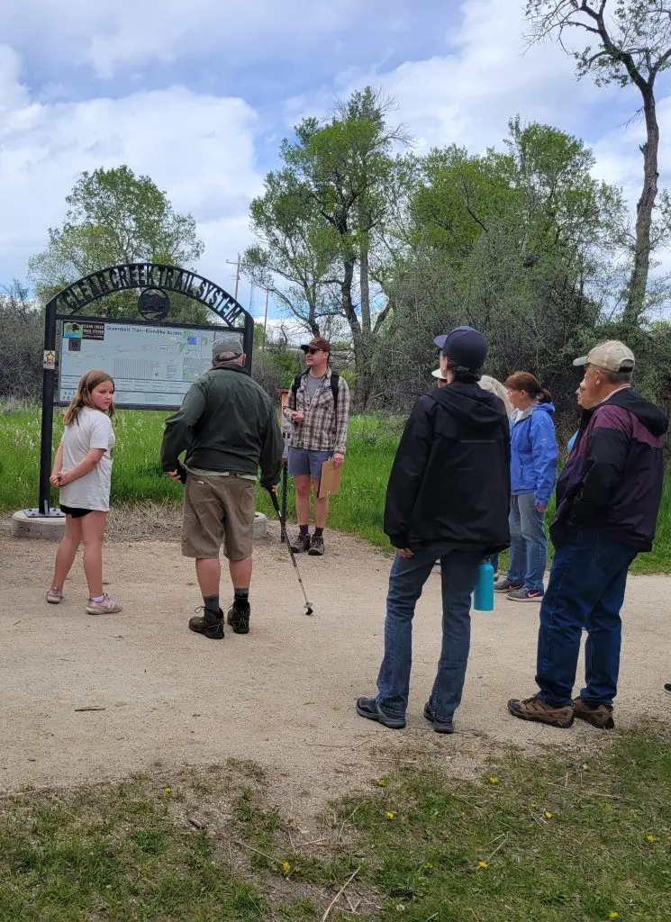group of people of varying ages stand in a circle around a man holding a clipboard beside a metal trailhead sign that says Clear Creek Trail System