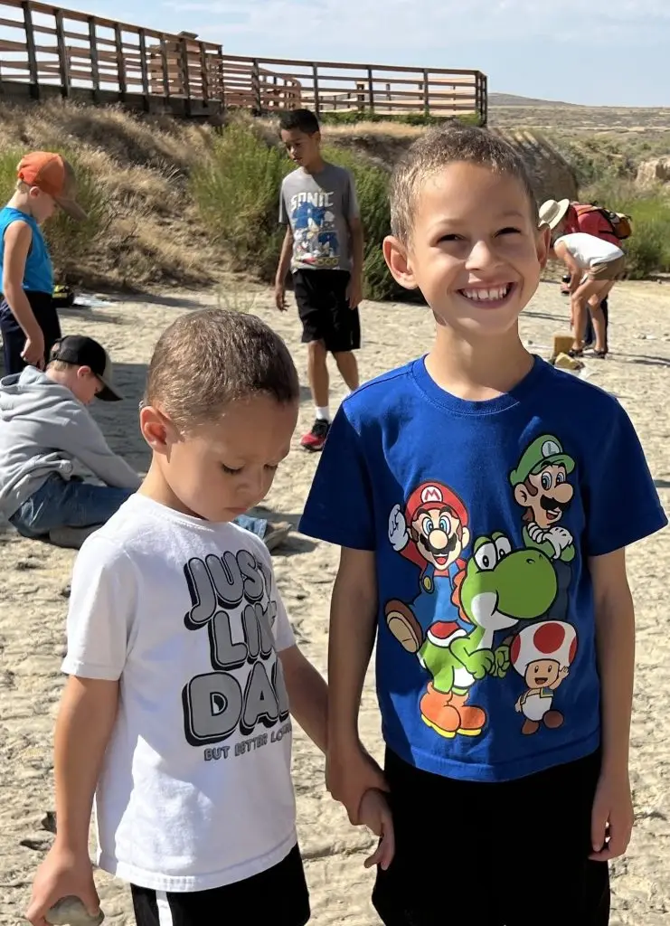 two young boys, one holding a rock and the other holding his hand, face the camera while other children and adults bend down to look at the rocky surface beneath their feet