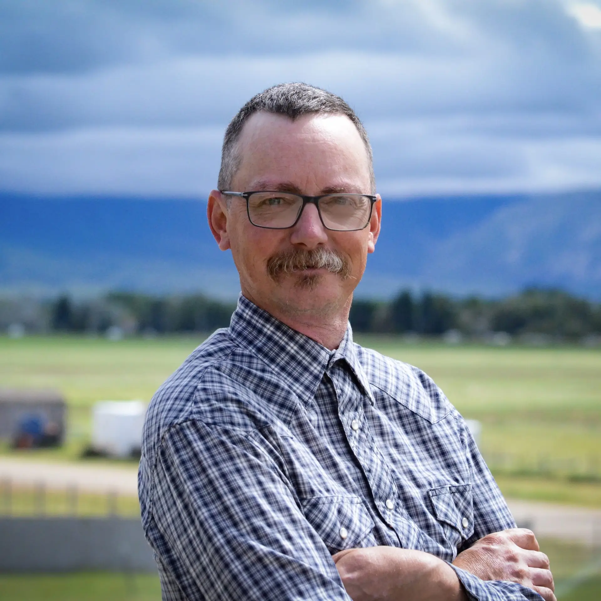 portrait of a man with glasses and arms crossed with green fields and mountains blurred in the background behind him