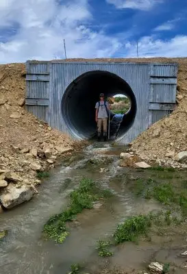 A young man stands in a culvert with a net. The culvert has a shallow stream of water flowing through it.