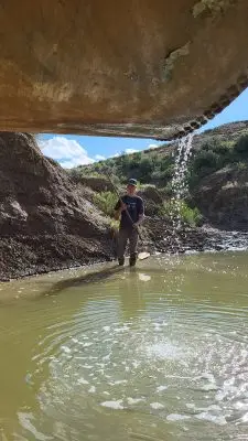 A young man stands in a pool of water with a long net. He is just beyond an overhang with water dripping off it.