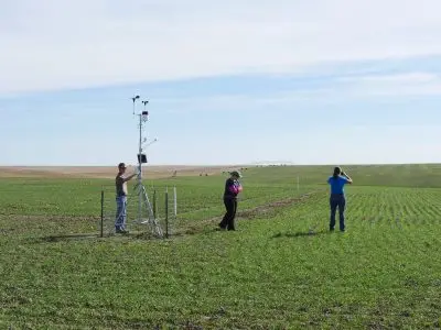 three people stand in a green field with pivot irrigation system in the distance. One man inspects a small weather station tower