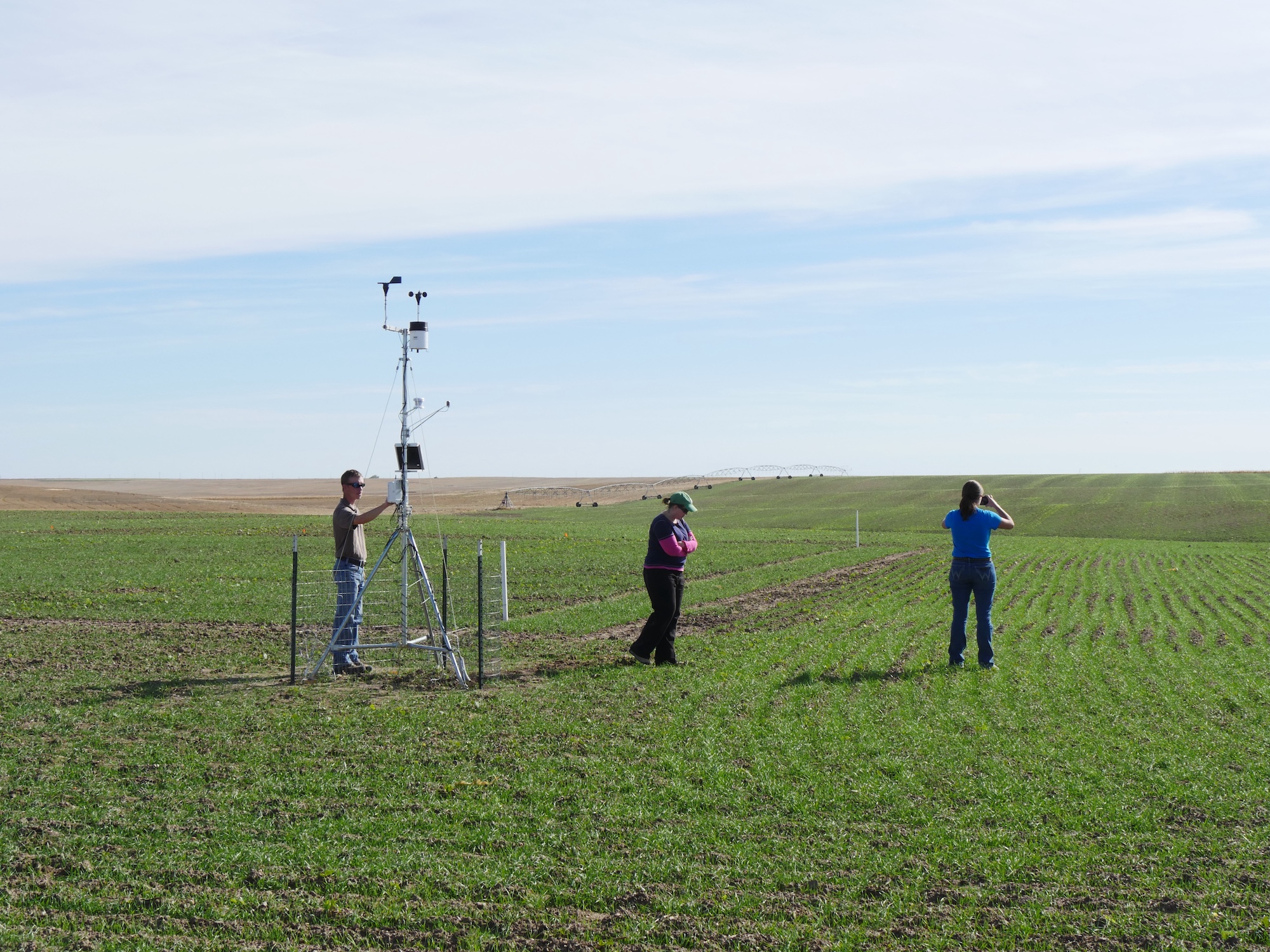 three people stand in a green field with pivot irrigation system in the distance. One man inspects a small weather station tower