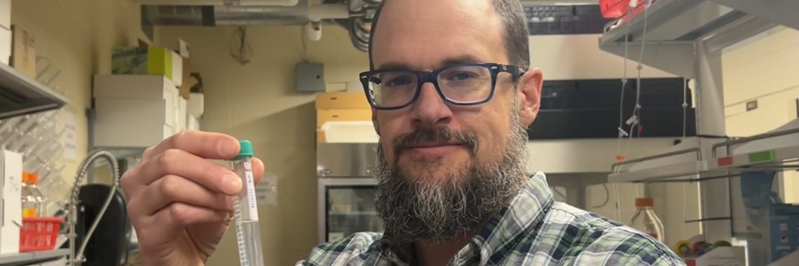 Smiling man holds up a test tube in two hands. The test tube contains a small beige blob in clear liquid