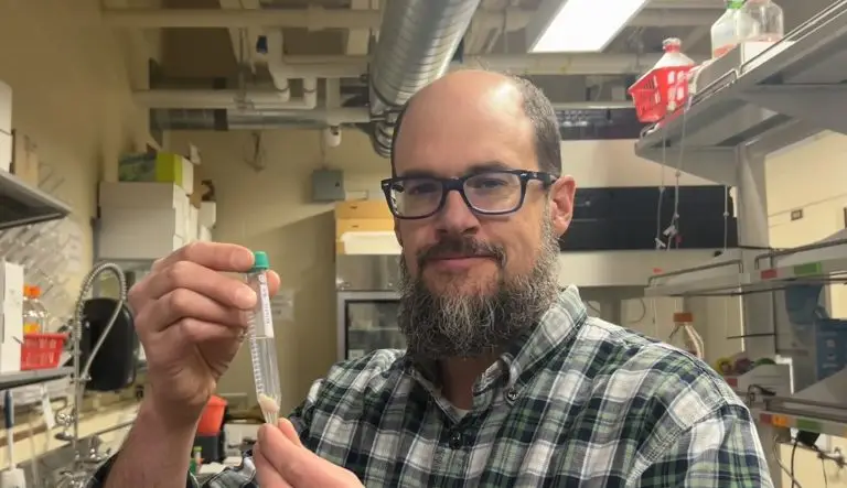 Smiling man holds up a test tube in two hands. The test tube contains a small beige blob in clear liquid