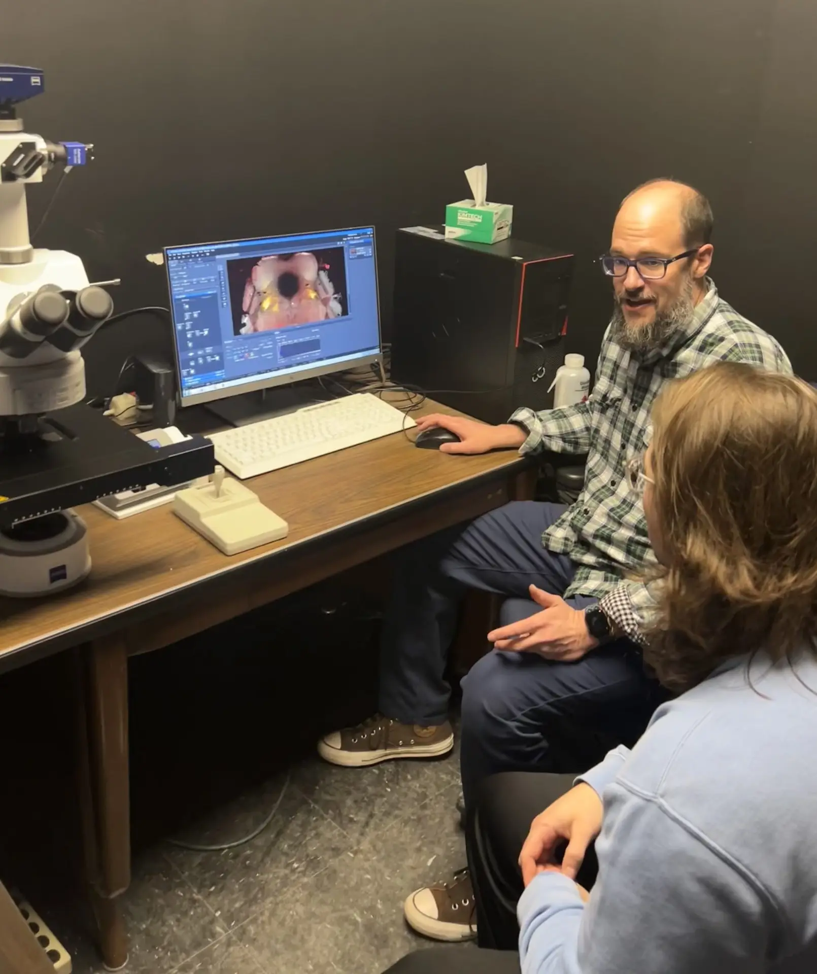 two scientists sit in front of a desk looking at a computer monitor showing a picture of a mouse brain