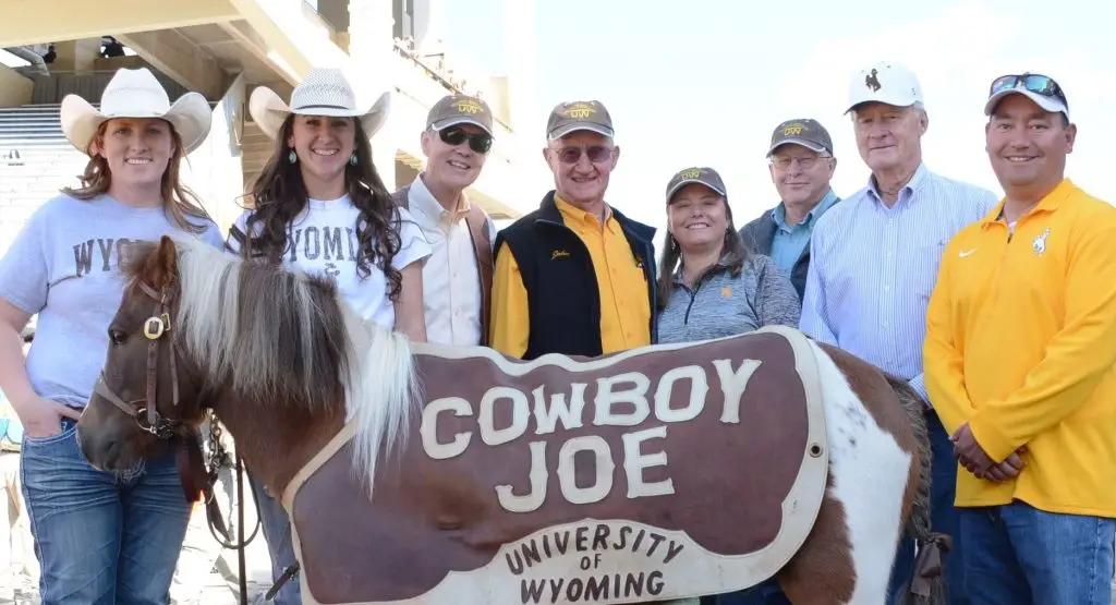 Eight smiling people wearing University of Wyoming gear stand together for a group photo with the UW mascot Cowboy Joe, a brown-and-white pony