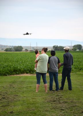 Several people watch a drone flying over a field.