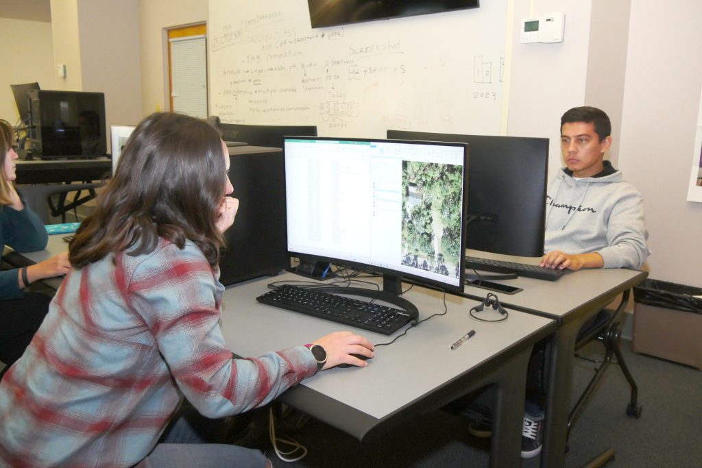 a man and woman sit opposite each other in a computer lab, each facing their computer monitor. Over the woman's shoulder, an aerial map and spreadsheet are visible.