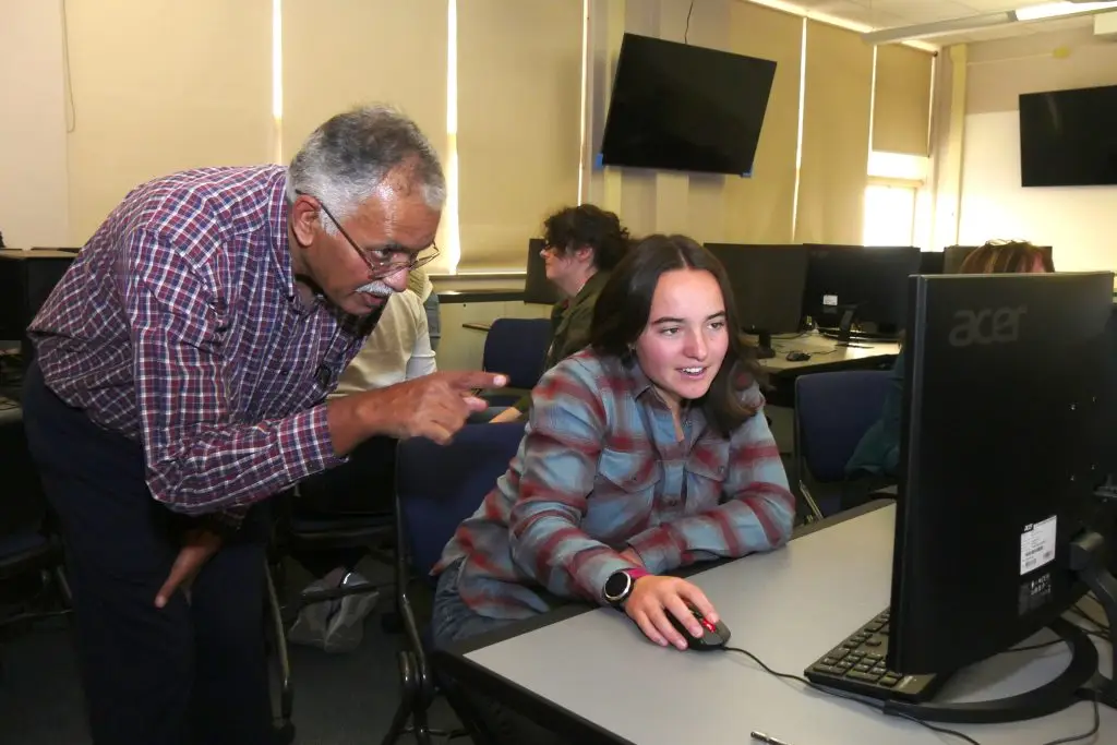 man bends down to look at a computer monitor with a seated woman with a hand on her computer mouse