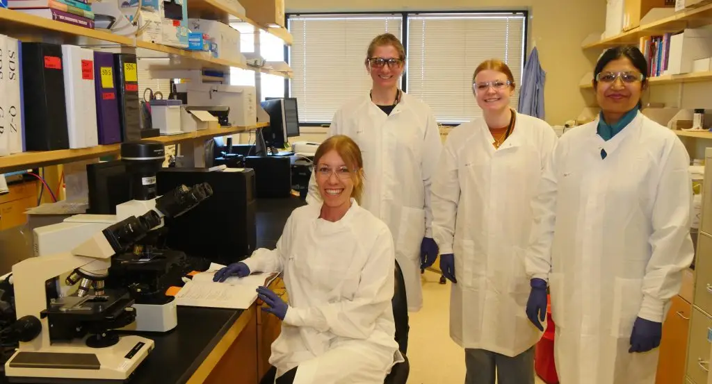 Four smiling female scientists wearing safety glasses, white lab coats, and purple gloves in an office space with two microscopes on a table and supplies stacked on wooden shelves