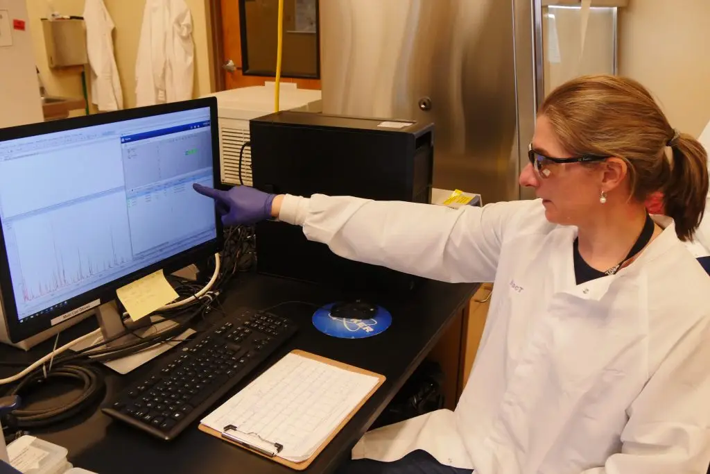 Woman wearing safety glasses and a white lab coat points a gloved hand at a computer monitor displaying a graph and data