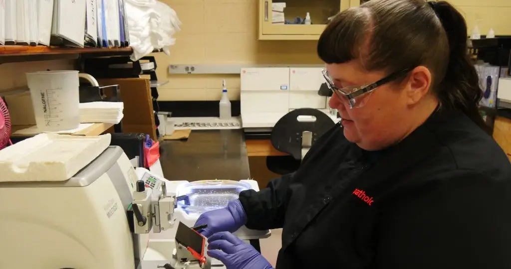 Woman wearing glasses, lab coat, and protective gloves operates a scientific device used cut thin slices of animal tissue samples for research