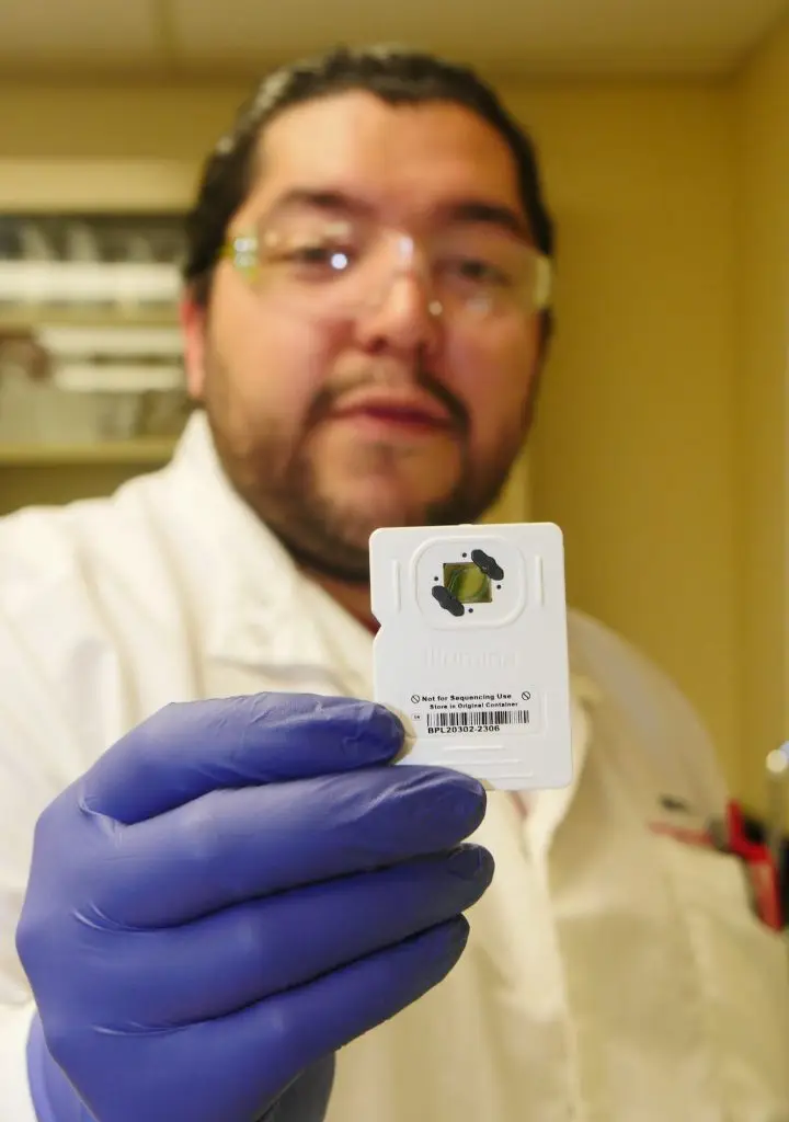 Man wearing lab coat and safety glasses holds out a gloved hand containing a small rectangular piece of equipment used in DNA sequencing