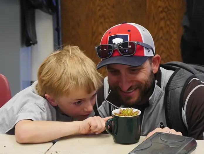 young boy and his father lean in close to look at a plant in a green ceramic mug on the table before them